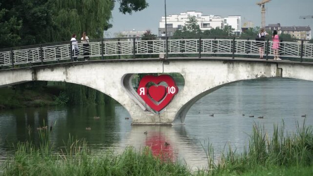 An old pedestrian bridge Ukrainian city, people looking distance, enjoying the view, ducks swimming lake, tall trees shore, a red heart installation in the middle bridge. Ivano-Frankivsk