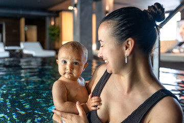 Baby in mothers arms at spa pool, direct gaze. Family wellness portrait.