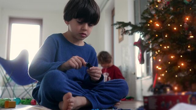 Child playing with toy pieces near Christmas tree with lights, holiday morning at home with family, joyful festive atmosphere filled with wonder and tradition