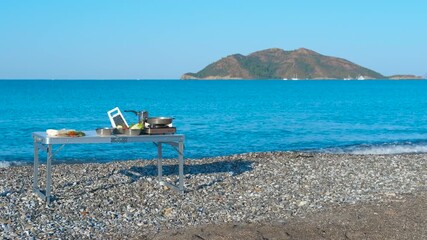 Camping table with food ready for cooking on a pebble beach. Portable camping table set with food on pebble beach, overlooking turquoise sea and distant island, creating outdoor dining atmosphere