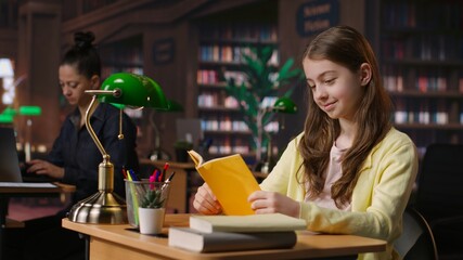 Young teen girl engrossed in reading books at the school library, focusing on literature and...