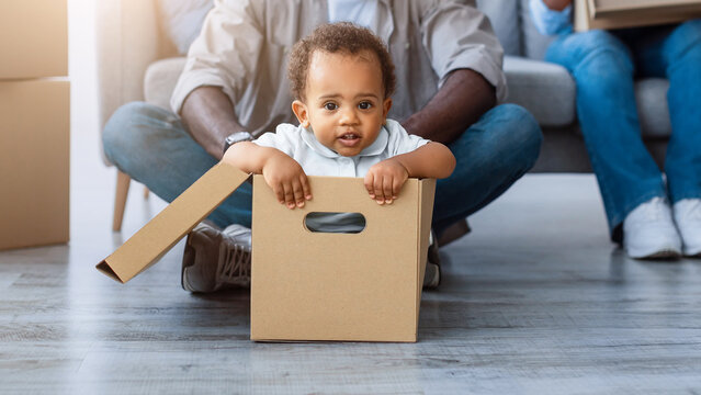 New Home, People And Real Estate Concept. Excited African American family having fun and celebrating moving day, cheerful father riding his little child in cardboard box container in living room - Powered by Adobe