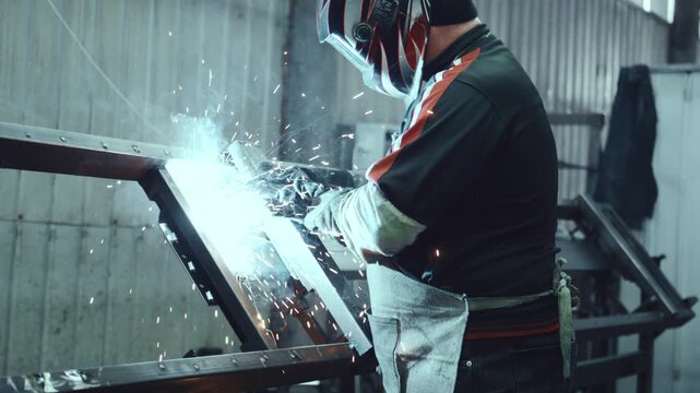 Industrial worker wearing a protective helmet welding a metal frame in a fabrication shop, with bright sparks and flashes illuminating the smoke filled workshop during the manufacturing process