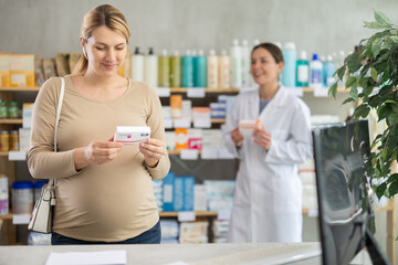 Fototapeta premium Pregnant woman choose pills of iodide and folic acid in pharmacy, examine packaging. Drug store staff offer supplements to customer in background. Health care, healthy lifestyle, prevention
