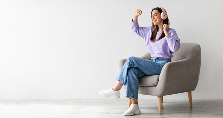 Cheery young lady in headphones listening to music and dancing while sitting in armchair against...