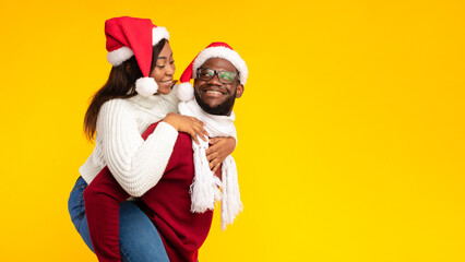 Xmas Concept. African American Husband Carrying Wife Piggyback Wearing Santa Hats On Christmas Posing Over Yellow Studio Background. Spouses Having Fun Celebrating New Year Holiday