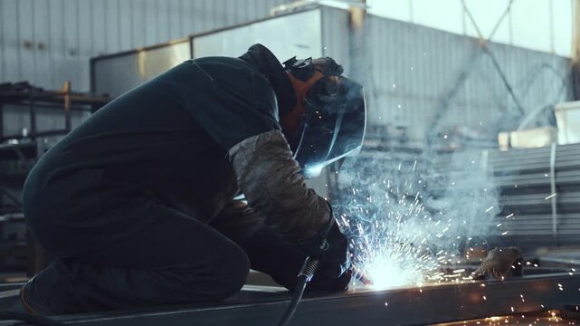 Industrial welder with a protective mask and gloves working with metal and creating bright sparks and smoke while welding in a heavy industry factory or a construction site workshop