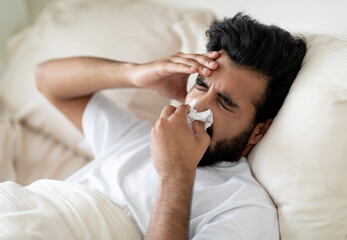 Closeup Shot Of Sick Indian Man Blowing Runny Nose In Napkin While Lying In Bed At Home, Ill Young Eastern Male Touching Head, Feeling Unwell While resting In Bedroom, Having Seasonal Flu Or Fever