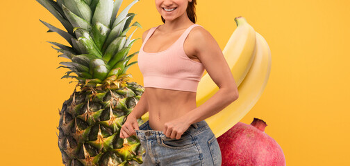 A cheerful young woman stands confidently with a smile, showcasing her weight loss by holding her...