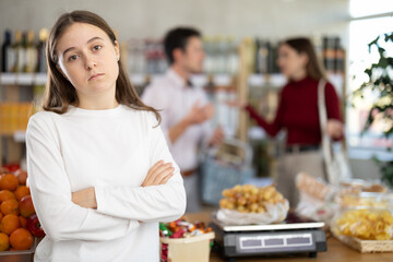 Sad teenage girl standing with arms crossed while parents argue in background at grocery store