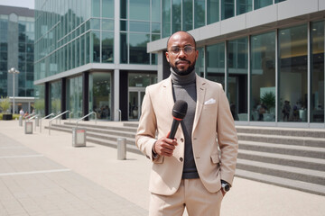 Portrait of young adult Black man standing outdoors in front of modern office building holding microphone, wearing glasses and suit, preparing for news report or interview