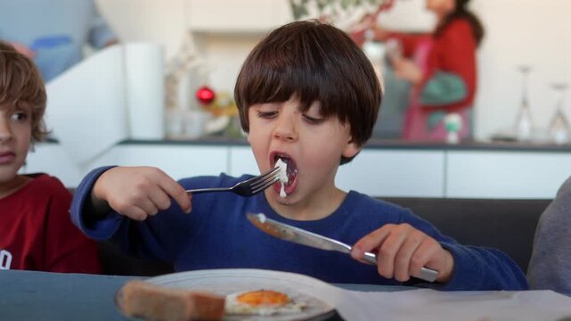Young boy chewing fried egg while holding fork and knife during breakfast with family at home kitchen table