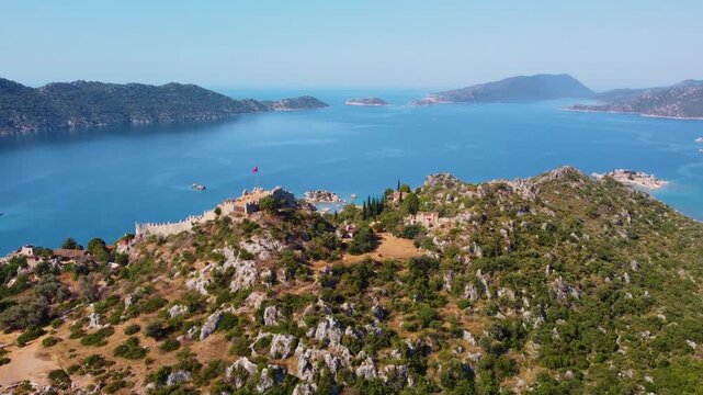 aerial footage showcasing the historic Simena Castle, also known as Kalek&ouml;y, perched on a hill overlooking the serene turquoise waters of Kekova Island and the Mediterranean Sea in Antalya Province, T