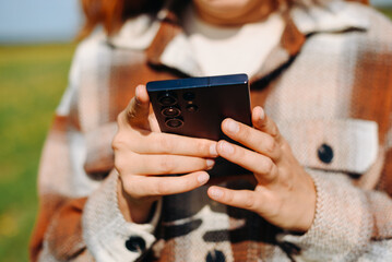 young woman holds smartphone in hands, standing on country road in green field in sunny summer day, beige plaid shirt, travel blogger, close-up view, no face, copy space