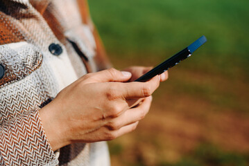 young woman holds smartphone in hands, standing on country road in green field in sunny summer day,...