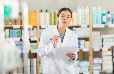 Skilled young female pharmacy employee in white coat standing with checklist, verifying product availability on shelves during routine inventory control