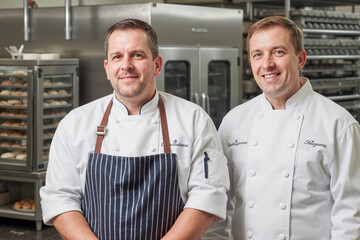 Two middle aged Caucasian men standing in commercial bakery kitchen, both wearing chef uniforms, smiling at camera, stainless steel ovens and racks with baked goods in background