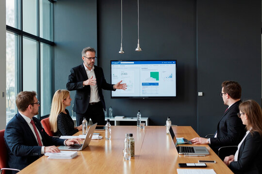 Caucasian middle aged man presenting data on screen to group of young adult and middle aged Caucasian men and women sitting at conference table using laptops and taking notes