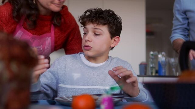 Father placing bread on table while mother kisses son on cheek, candid family scene showing everyday affection and warmth during mealtime at home