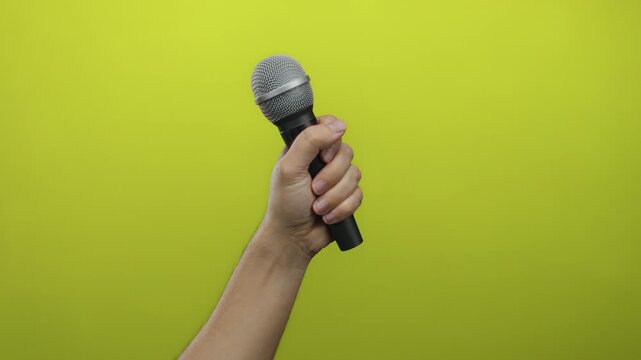 Man holding microphone against vibrant yellow wall, showcasing male hand gripping mic with green background, conveying performance or speech concept in isolated setting.