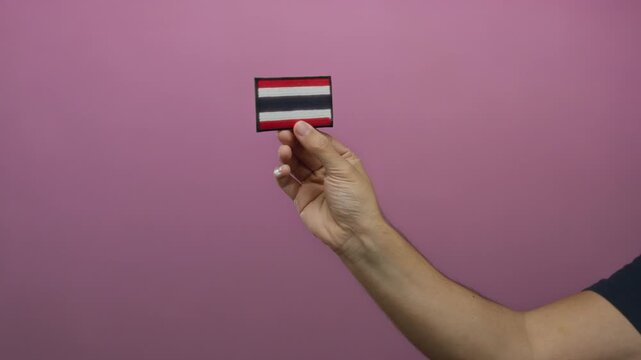 Man holding embroidered thailand flag patch with hand against pink isolated background implying cultural focus and national identity.