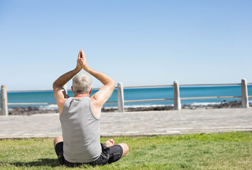 Senior man wearing workout gear practicing yoga on grassy lawn by paved walkway and seaside railing