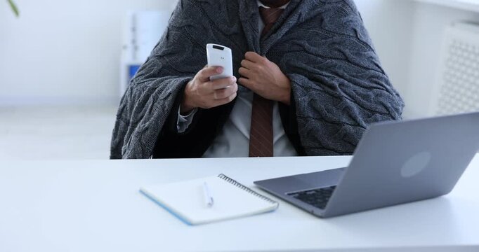 Man with blanket suffering from cold temperature and using remote to control air conditioner in office, closeup