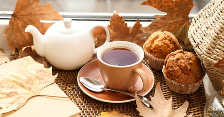 Teapot with cup of hot beverage, tasty cupcakes, book and autumn leaves on windowsill in room