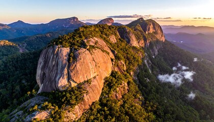 Aerial view of a lush mountain range at sunset, featuring rocky peaks, dense forest, and warm golden light