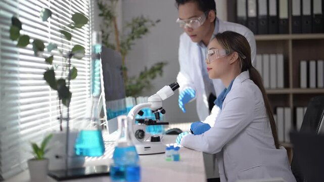 Two scientists are working together in a lab, one of them pointing at a computer monitor. The lab is filled with various scientific equipment and supplies, including a large number of bottles