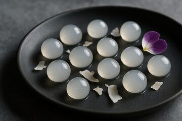 Delicate translucent mochi dessert balls on black plate with edible flower petal and coconut flakes, modern minimalist presentation