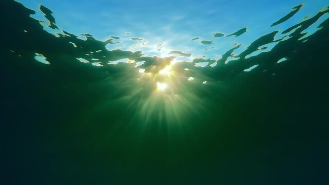 Sun beams playing with brightly flashes beneath surface with waves at golden hour. Slow-motion: vibrant underwater sunrays shine below water, illuminating sea depths, backlit by brilliant sunburst
