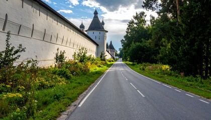 An open road, flanked by a long, aged white wall on the left, and a vibrant green forest on the right, stretching towards the distant sky