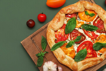 Wooden board of tasty tomato galette with basil and garlic on green background