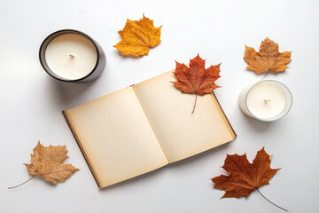 Minimal autumn composition with candles, dried maple leaves, a knitted scarf, and a vintage notebook on a white surface.