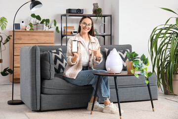 Young woman with blank mobile phone and air humidifier on table at home