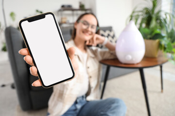 Young woman with blank mobile phone and air humidifier at home, closeup