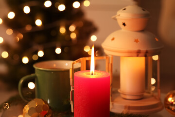 Burning candle, lantern, cup of tea and decorations against Christmas tree indoors, closeup