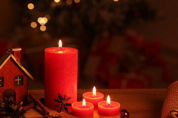 Composition with burning candles, spices and house figure on wooden table against Christmas tree indoors