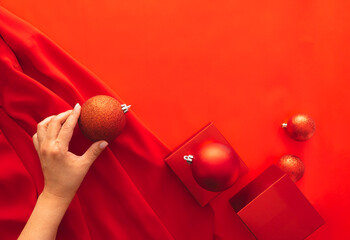 A hand places a glittering orange Christmas ornament on a red fabric surface. Nearby are red gift...