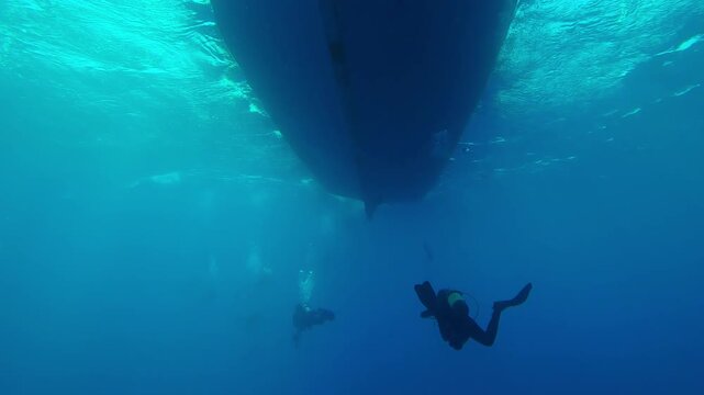 Back view of group of scuba divers swimming in turquoise water towards diving boat on sunny day in backlit, Slow motion of divers returning underwater to diving safari boat