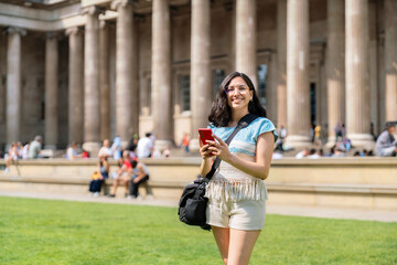 Young smiling Latina woman outside the British Museum in London, England, using her mobile phone. She wears a woven blouse, beige shorts. People sit and enjoy the green garden behind her