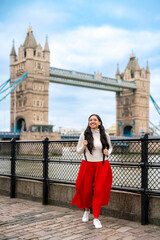 Young happy Latina woman stands along the Thames in front of the iconic Tower Bridge in London, England. Dressed turtleneck and skirt, she enjoying the scenic view and tourist vibe near the landmark