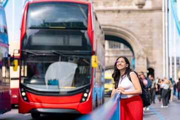 Naklejka premium Happy young Latina woman smiles beside a red double-decker bus on Tower Bridge in London, England. Wearing turtleneck, red skirt and backpack, she enjoys city life and British landmarks on a sunny day