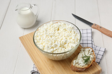 Glass bowl of fresh cottage cheese with bread slice and jug of milk on white wooden background
