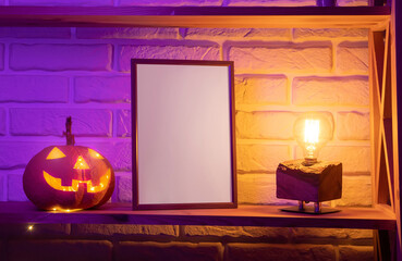 A picture frame with a white blank sheet of paper mockup with space for design and text stands on a wooden shelf next to a scary Halloween pumpkin lantern in a dark evening room with purple lighting.