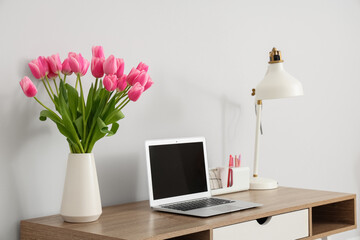 Beautiful bouquet of tulip flowers with laptop and lamp on table at workplace