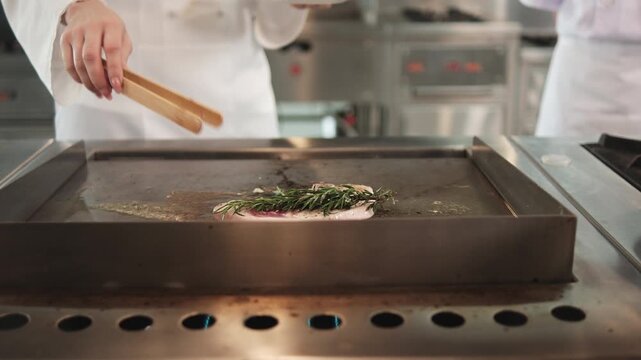 Close-up shot of chef hand-roasting a beef steak with tongs in frying pan, herbs and spices cooking ingredients in restaurant kitchen, professional in gourmet foods and delicious meal occupation.  