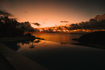 A stunning infinity pool reflects the vivid orange and red colors of a dramatic ocean sunset,...