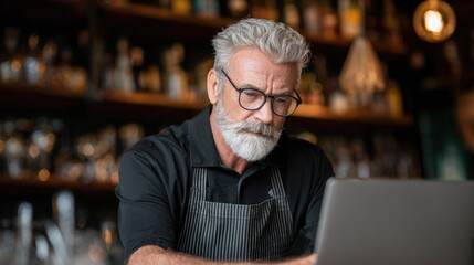 A man with gray hair and beard wearing glasses, working at a bar, using a laptop.